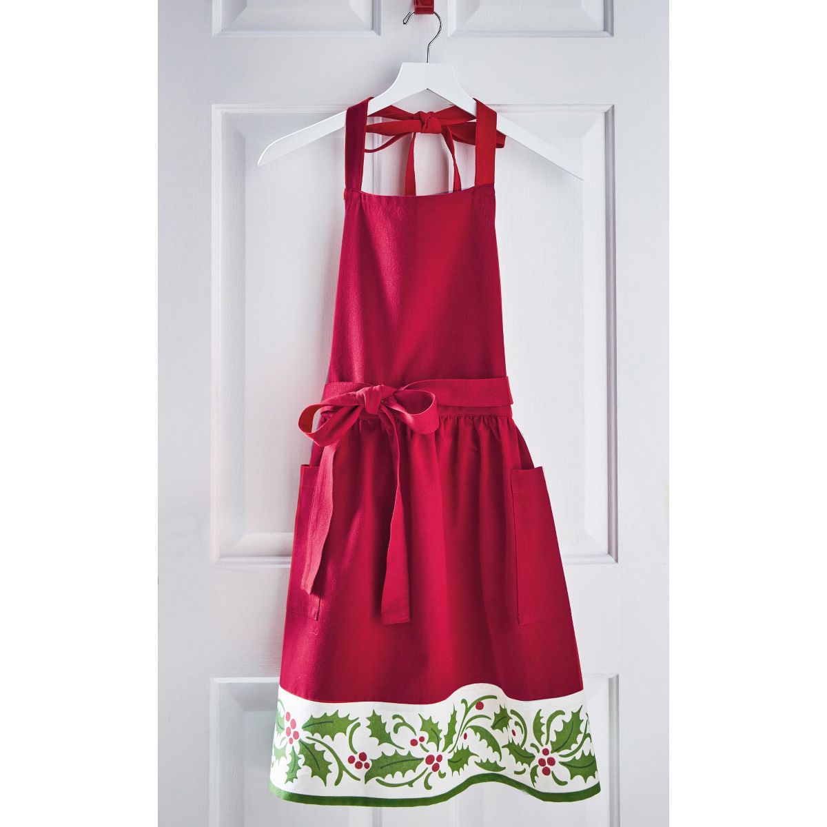 Red Christmas apron with holly berry border print and tied waist bow on hanger