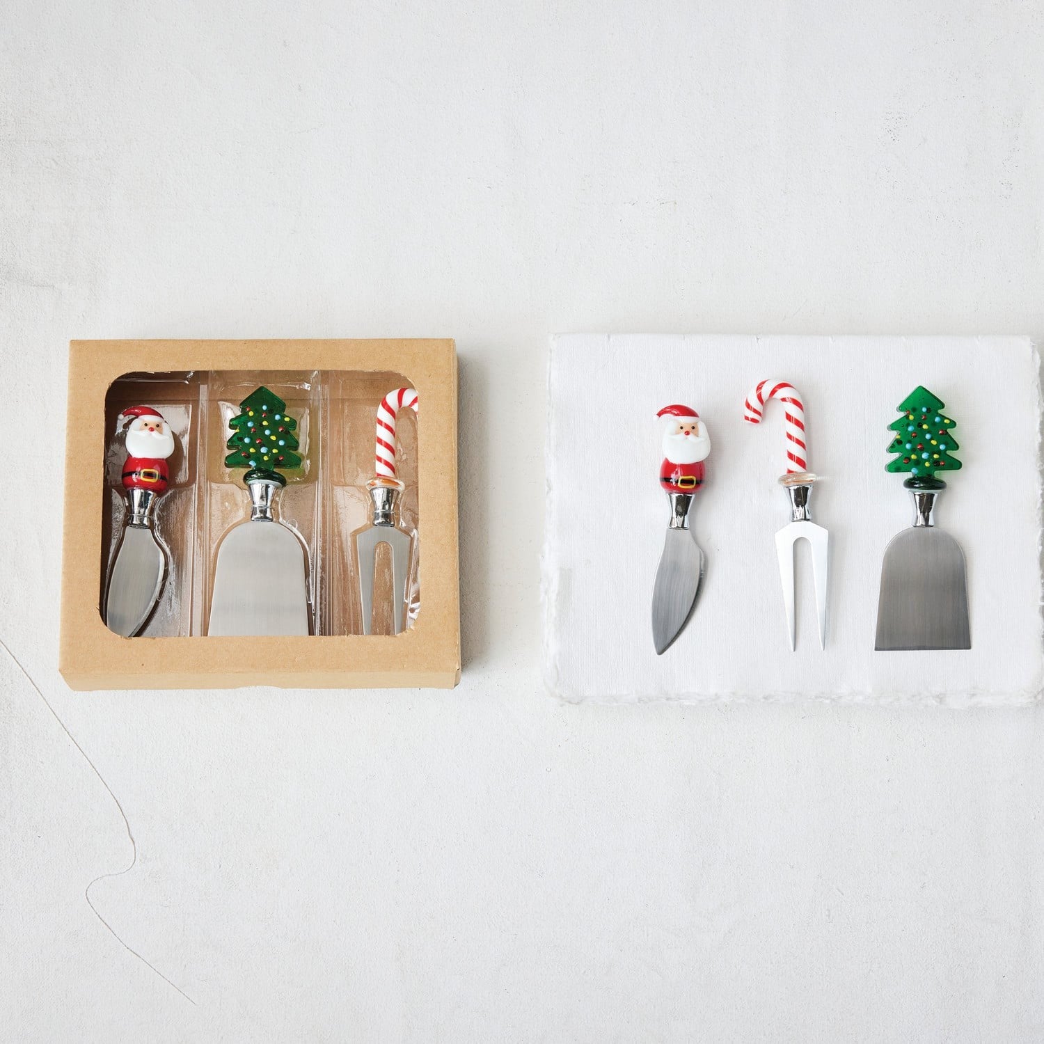 Holiday cheese knives set of 3 with Santa, candy cane, and Christmas tree handles, shown in kraft gift box and styled on m...