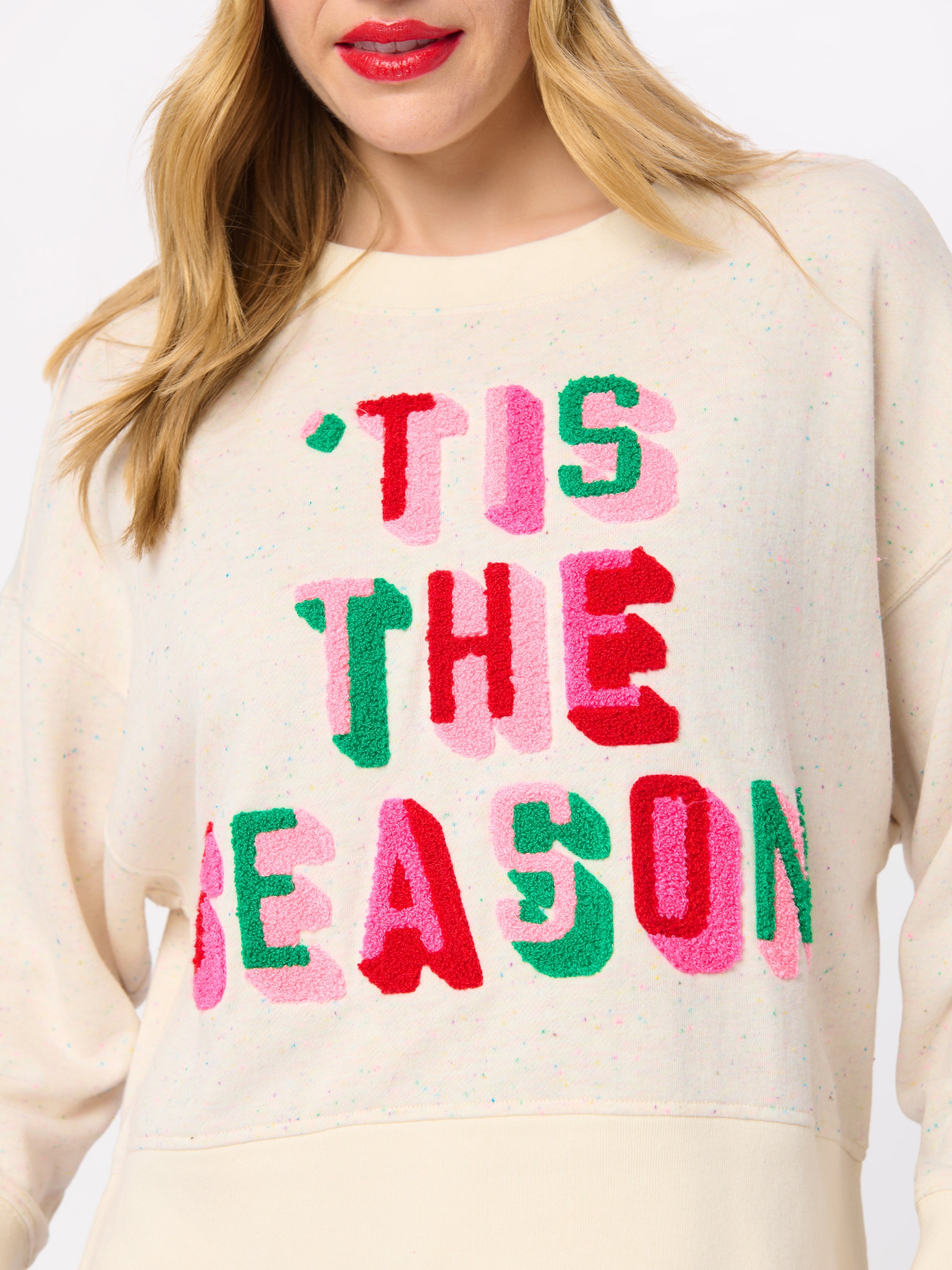 Close-up detail of cream sweatshirt with colorful 'Tis The Season embroidered text in pink, green, and red