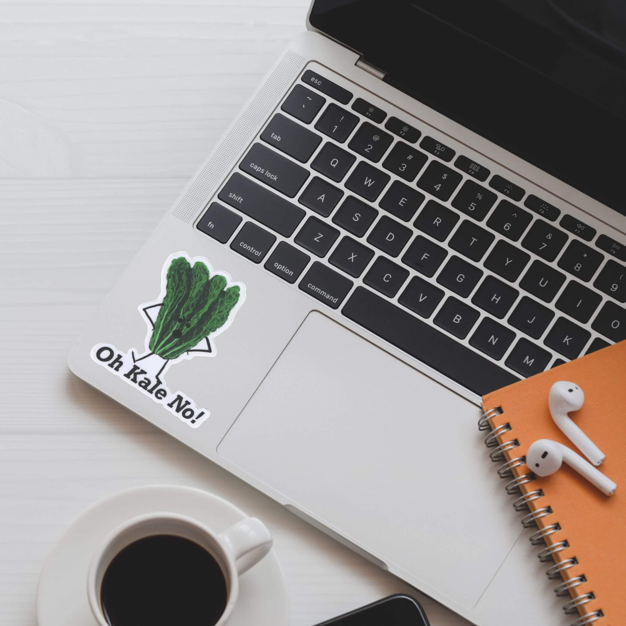 Oh Kale No sticker displayed on a MacBook laptop beside coffee and notebook